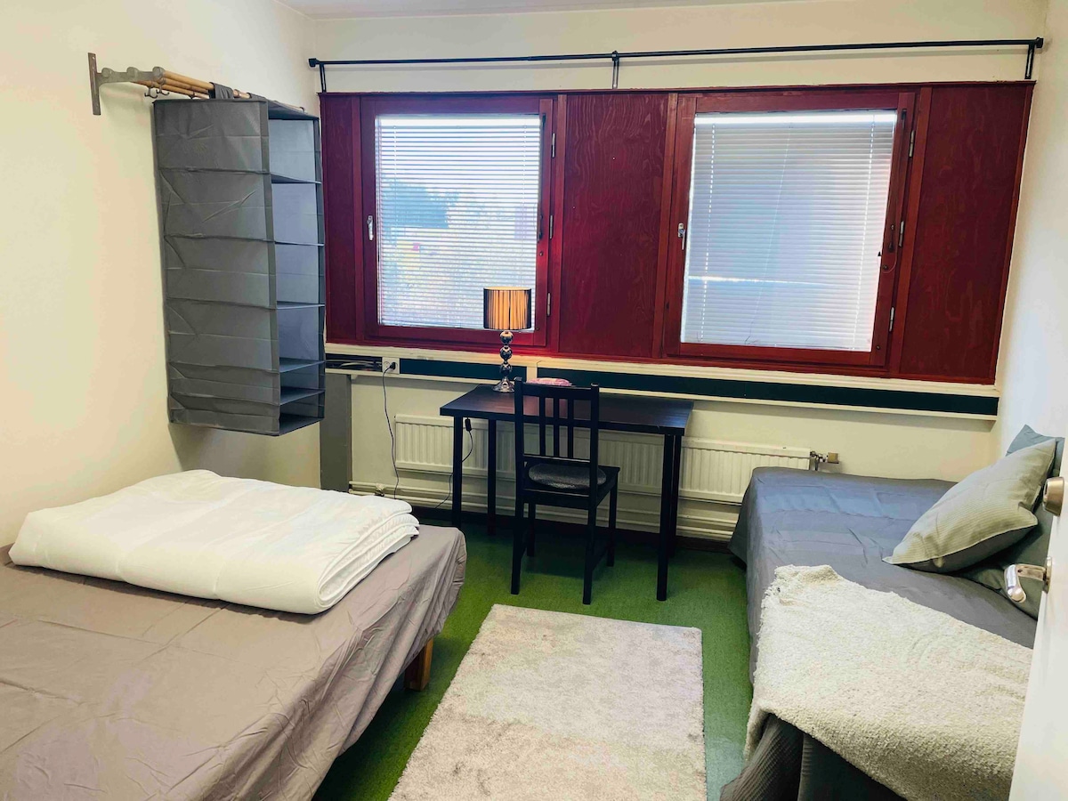 A simple sleeping area features two beds with light bedding, complemented by a desk and chair. A lamp sits on the desk, while large windows dressed with blinds allow natural light into the space. Neutral-colored walls enhance the room's functionality.