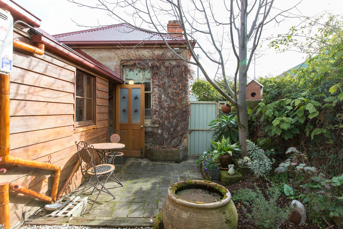 A private courtyard features natural stone paving and a variety of plants, including a tree, surrounded by stone and timber walls. A small round table with two chairs is positioned near the entrance, inviting outdoor relaxation.