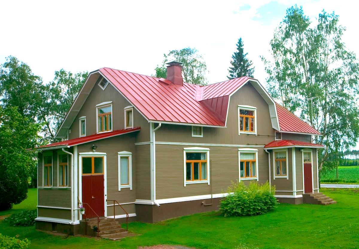 A two-story house is depicted, featuring a gray exterior with white trim and a red metal roof. Multiple windows provide natural light, while a porch and steps lead to the entrance. Lush greenery surrounds the property, creating a peaceful outdoor environment.