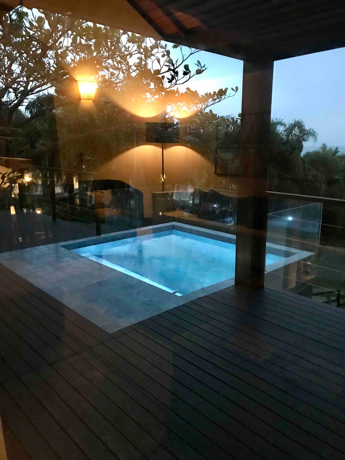 A private pool is positioned on a wooden deck, reflecting soft lights from nearby lanterns. Surrounding greenery adds an element of nature to the tranquil setting, while a clear view of the sky indicates dusk.