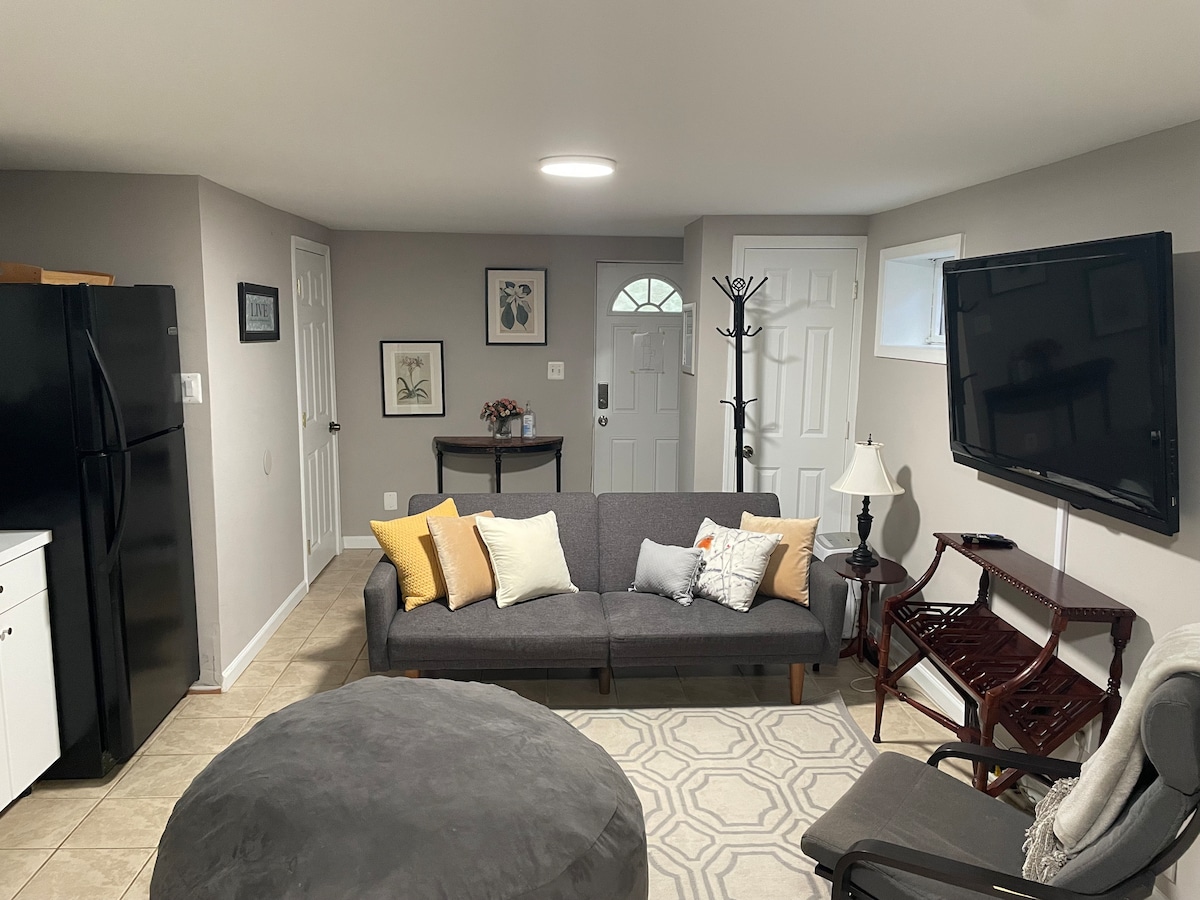 A spacious living area is furnished with a gray futon adorned with decorative pillows. A coffee table and a large, soft ottoman complement the seating. A television is mounted on the wall, and light enters through several windows, illuminating the neutral-toned decor.