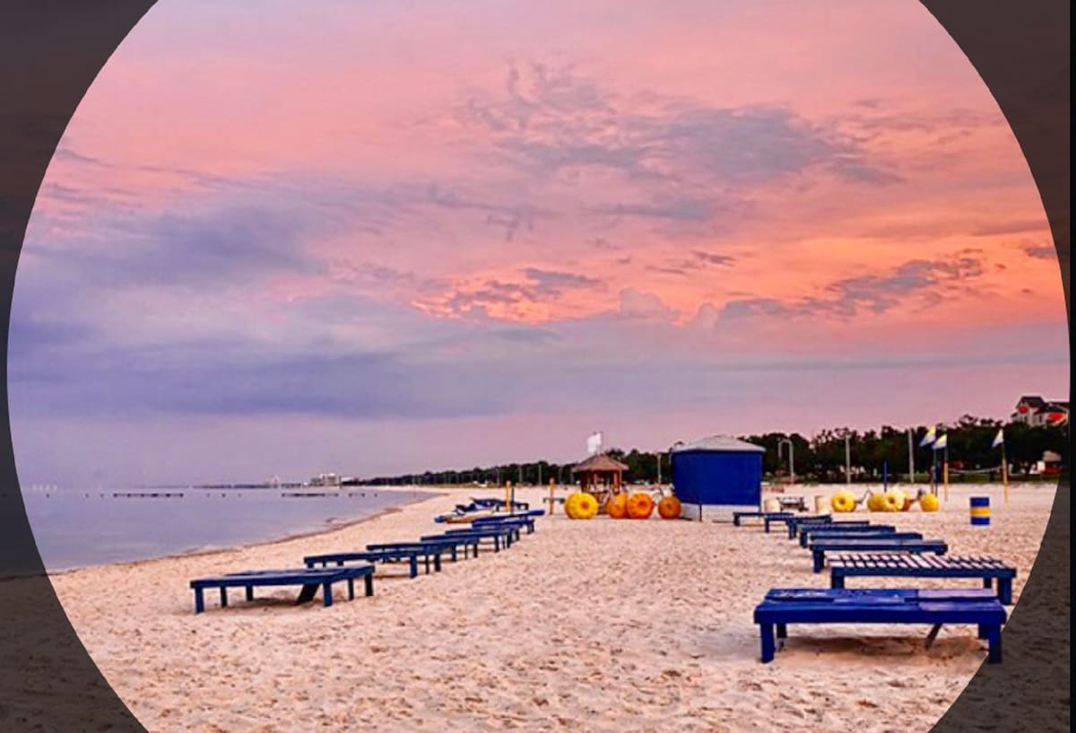 A beach scene is displayed, featuring rows of wooden lounge chairs on soft sand. The horizon is graced with a colorful sunset, casting a warm glow over the serene waters. Brightly colored inflatable rings are arranged nearby, inviting leisurely water activities.