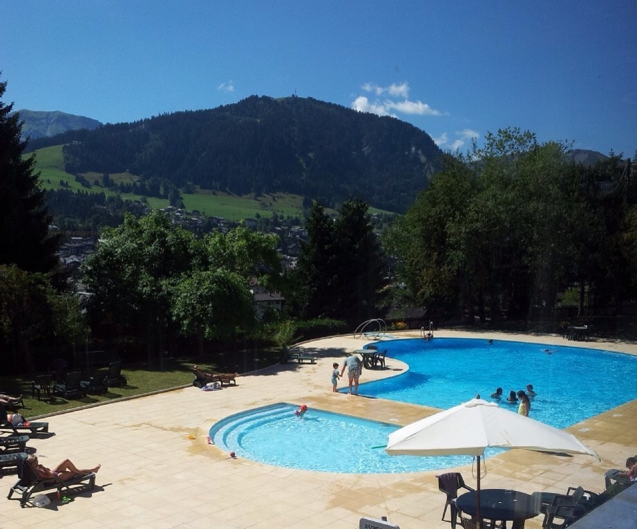 An outdoor swimming pool is surrounded by chaise lounges and shaded by large umbrellas. The inviting blue water reflects the sun amidst a scenic mountain backdrop and green landscape. Guests can be seen enjoying the space while trees provide additional greenery around the area.