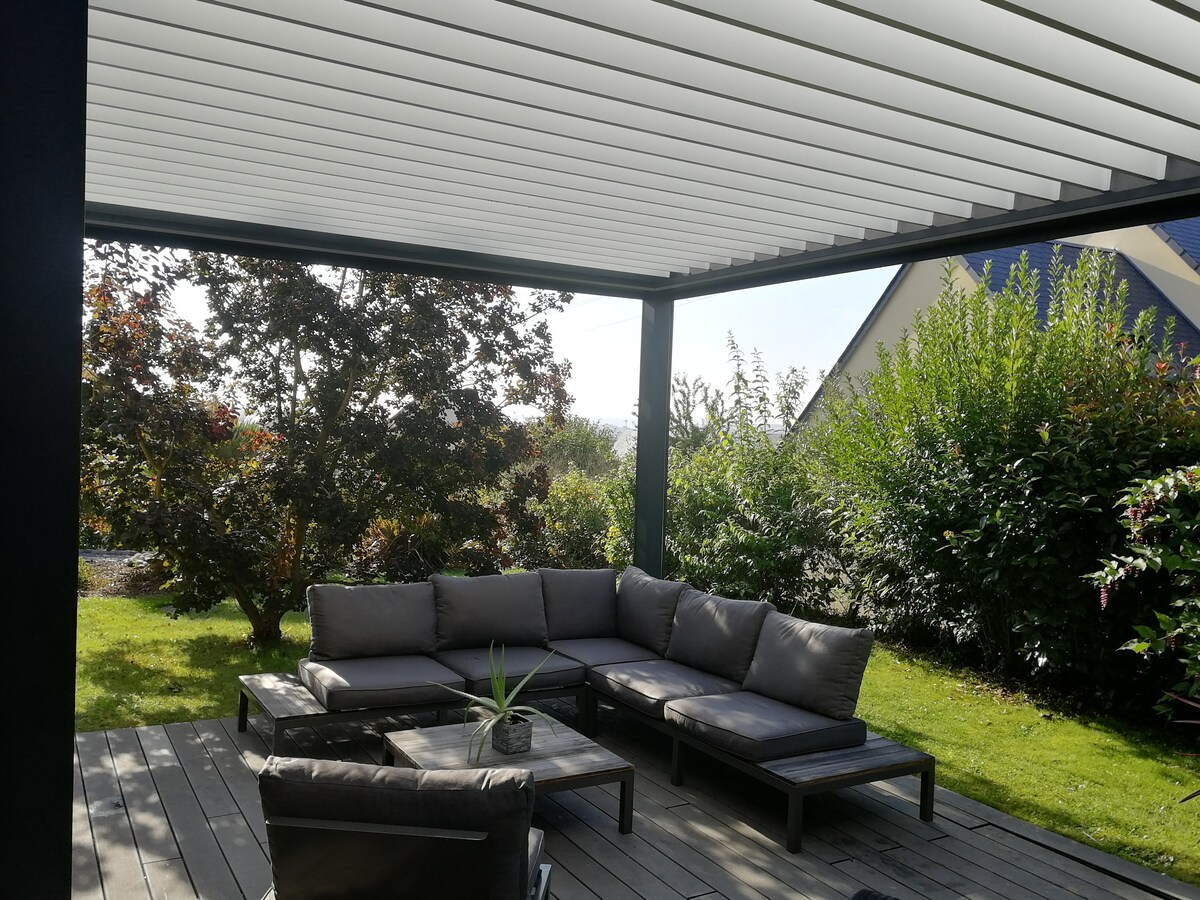 An outdoor seating area is presented under a modern pergola, featuring a dark sectional sofa with plush cushions. A coffee table is placed in the center, surrounded by lush greenery, including shrubs and trees. The wooden deck provides a natural setting for relaxation.