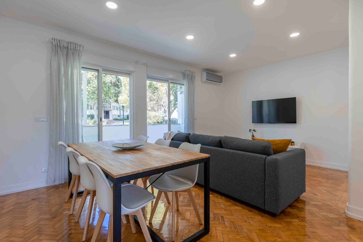 A spacious living and dining area features large windows allowing natural light to illuminate the room. A grey sofa is positioned near a mounted television. A wooden dining table with white chairs is visible, offering an inviting space for meals or gatherings.