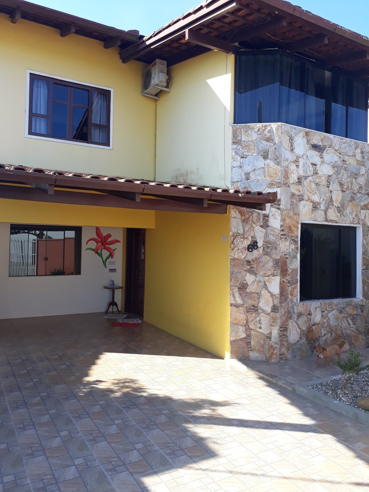A two-story house features a stone accent wall and large windows that allow natural light to enter. A vibrant yellow exterior complements the surrounding area, with a welcoming entrance that includes a decorative plant and a shaded porch area.