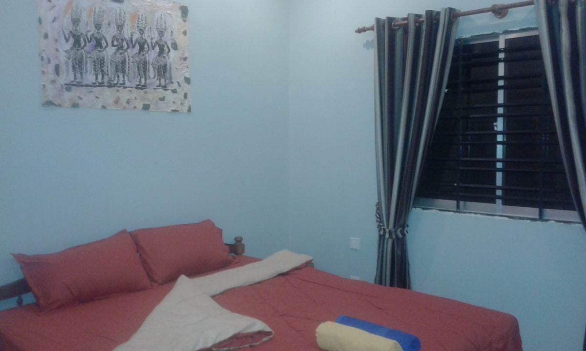 A cozy room features a spacious bed with red linens and two pillows. A light blanket is draped across the bed. The walls are painted light blue, with an artistic wall hanging above the bed. Blackout curtains frame the window, complementing the room's decor.