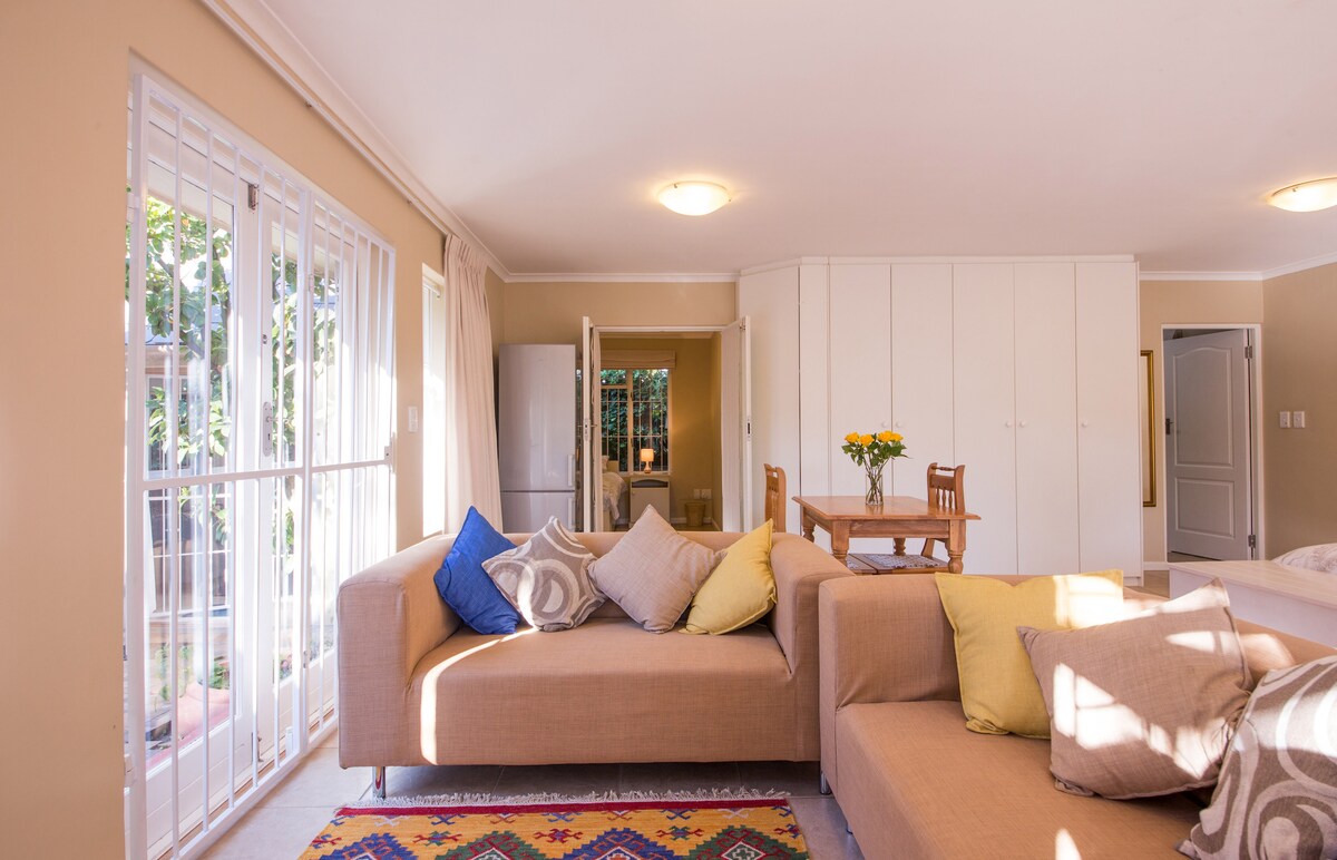 A light-filled living area features a cozy sectional sofa adorned with colorful cushions. A wooden dining table is positioned nearby, complemented by a vase of yellow flowers. Large windows allow natural light to brighten the space, showcasing the inviting decor.