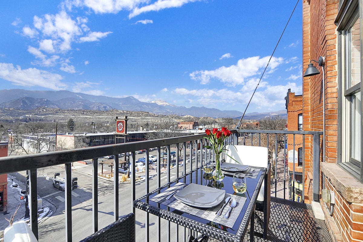 A balcony dining area features a table set for two, accompanied by a vase of red roses. Scenic views of Pikes Peak and surrounding landscapes are visible in the background, with a clear blue sky enhancing the outdoor setting.