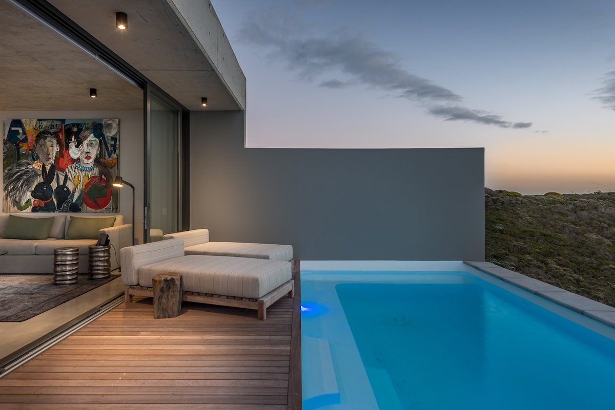 An outdoor area features a minimalist lounge set overlooking a pool with soft lighting. The adjacent indoor space has a vibrant artwork displayed and large glass doors that open to the deck, enhancing the seamless connection between indoor and outdoor environments.