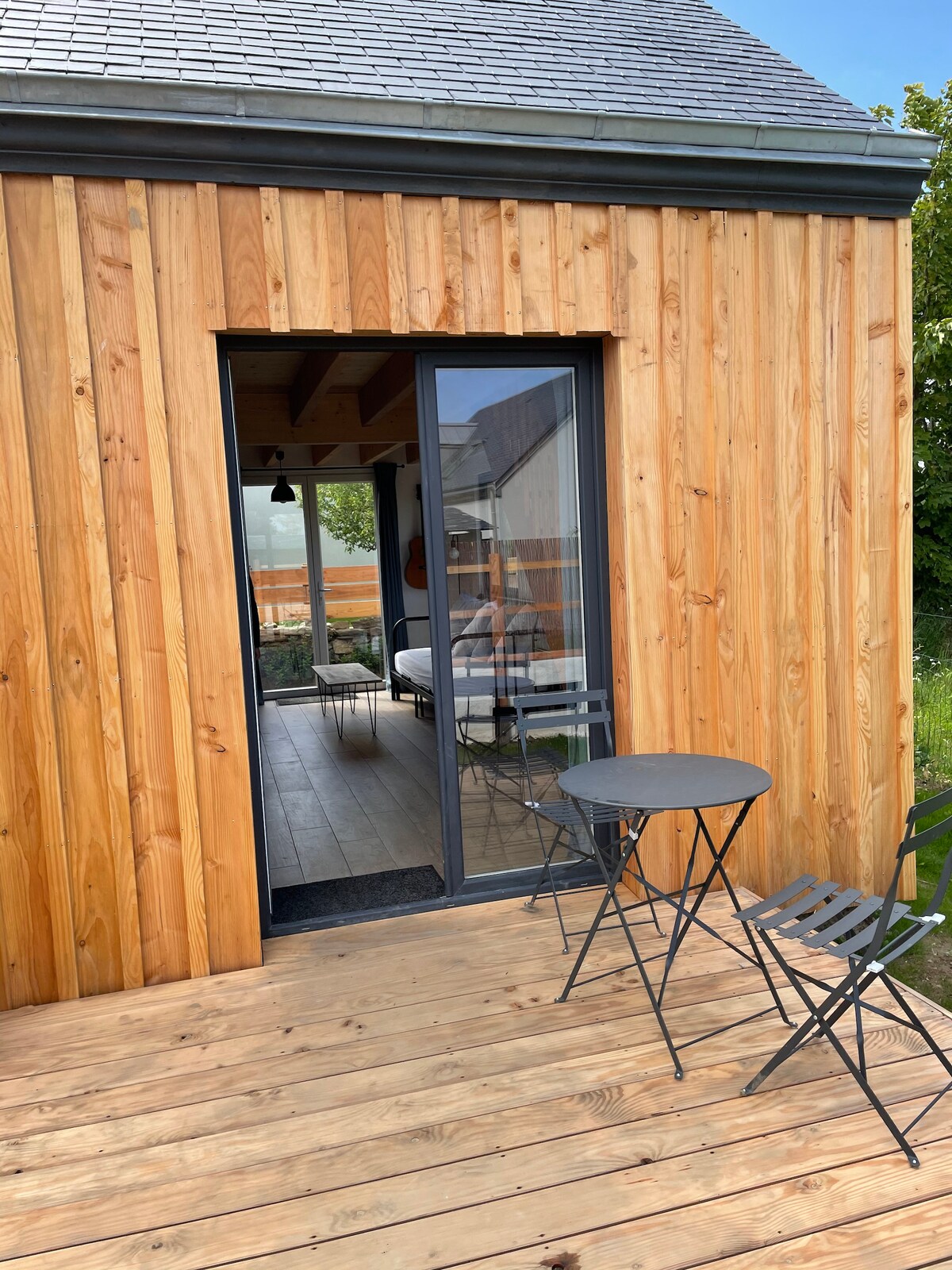 A wooden exterior features a sleek glass door leading to the interior space. A small round table and two folding chairs are positioned on the terrace, ideal for enjoying the outdoor setting. The surrounding greenery adds a natural touch to the ambiance.