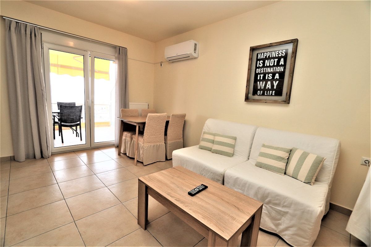 A light-filled living space features a comfortable sofa and a rustic wooden coffee table. A dining area with a built-in table and black chairs is visible in the background. Light curtains frame sliding glass doors that lead to a balcony, enhancing the airy atmosphere.