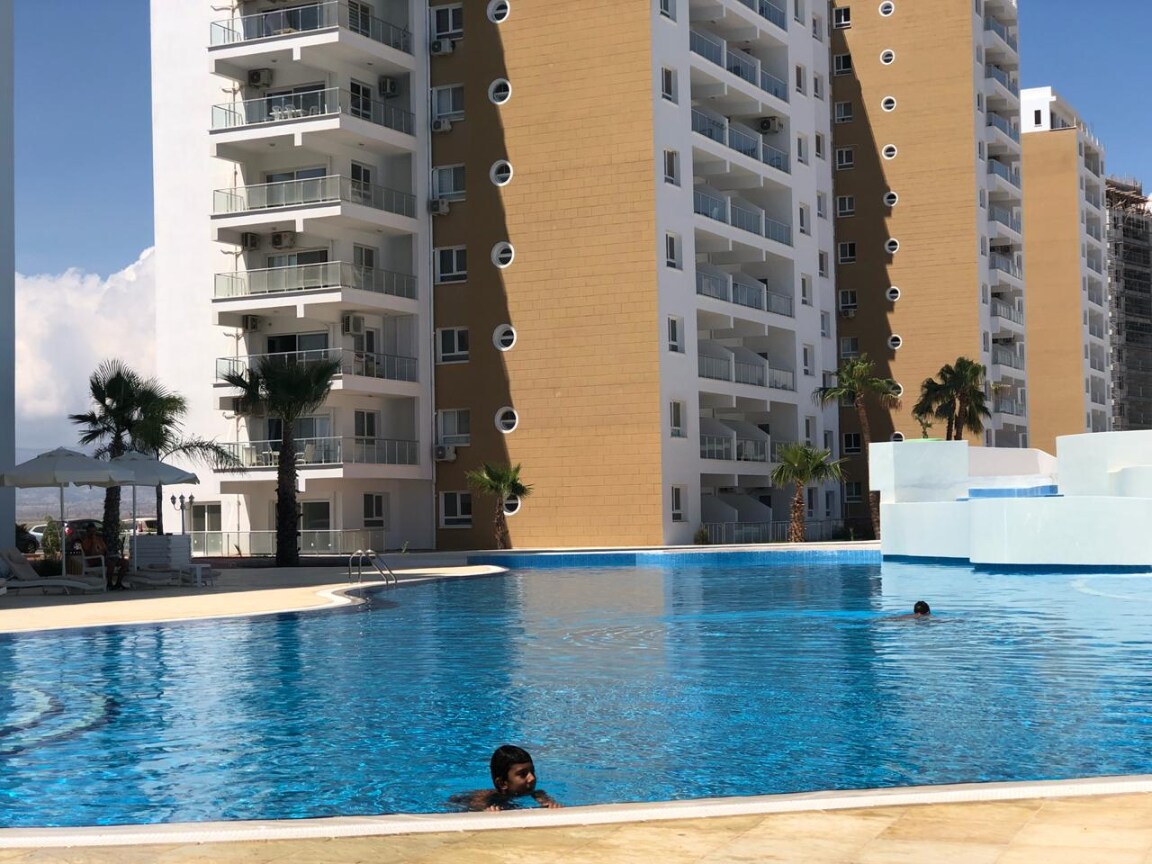 A large, inviting swimming pool is surrounded by modern residential buildings. The pool features a calm, clear blue surface reflecting the sunny sky. Palm trees line the edges, adding to the relaxing atmosphere, while a few guests enjoy the water.