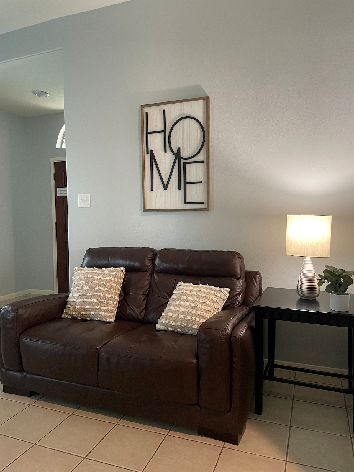 A cozy seating area features a brown loveseat adorned with two decorative cushions. A small side table holds a lamp and a potted plant. A framed wall art piece with the word 'HOME' adds a personal touch to the space.