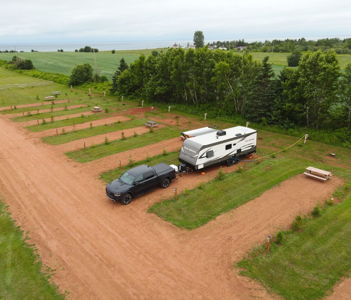 An overhead view of a spacious trailer park shows several designated spots equipped with power, water, and sewer connections. A black truck is parked next to a camper. Picnic tables are positioned throughout the grassy area, framed by a backdrop of trees and the ocean in the distance.