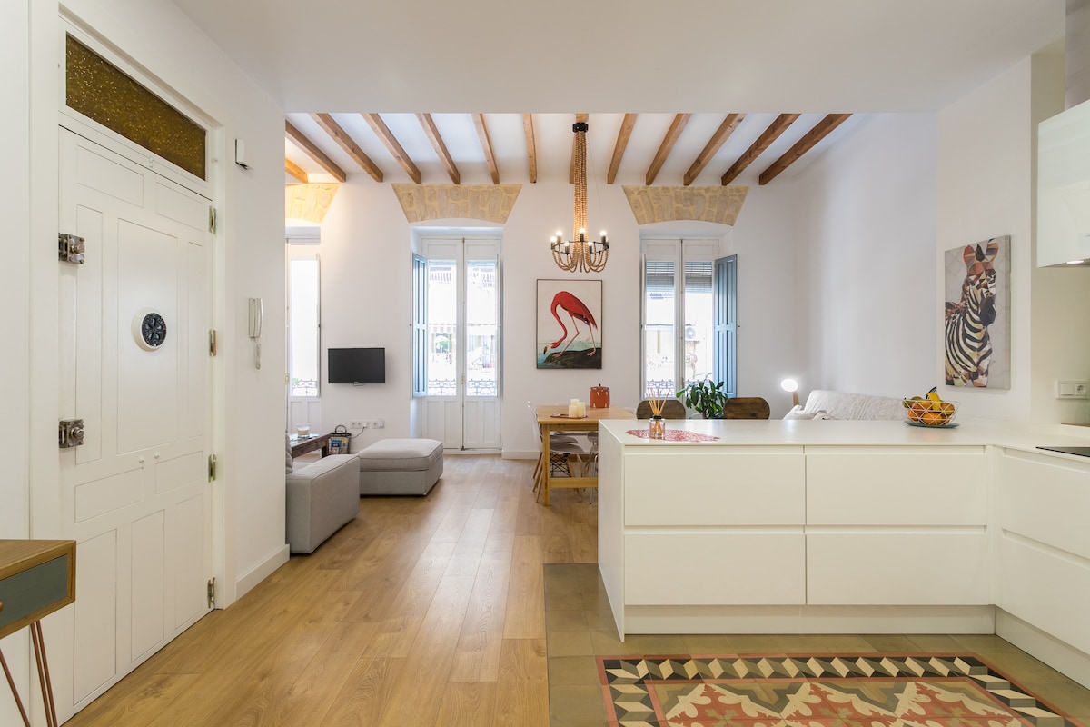 An open living space features wooden beams overhead and stylish furnishings. A modern kitchen area is complemented by a dining table and a comfortable seating area, while large windows allow natural light to fill the room. Colorful artwork adds a touch of personality.