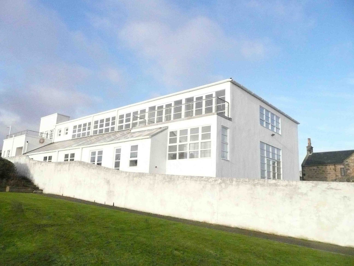 The building is characterized by its large white exterior and numerous windows, allowing natural light to fill the interior. The surrounding green lawn adds to the spaciousness of the property, while a stone wall provides subtle delineation from the outdoor area.