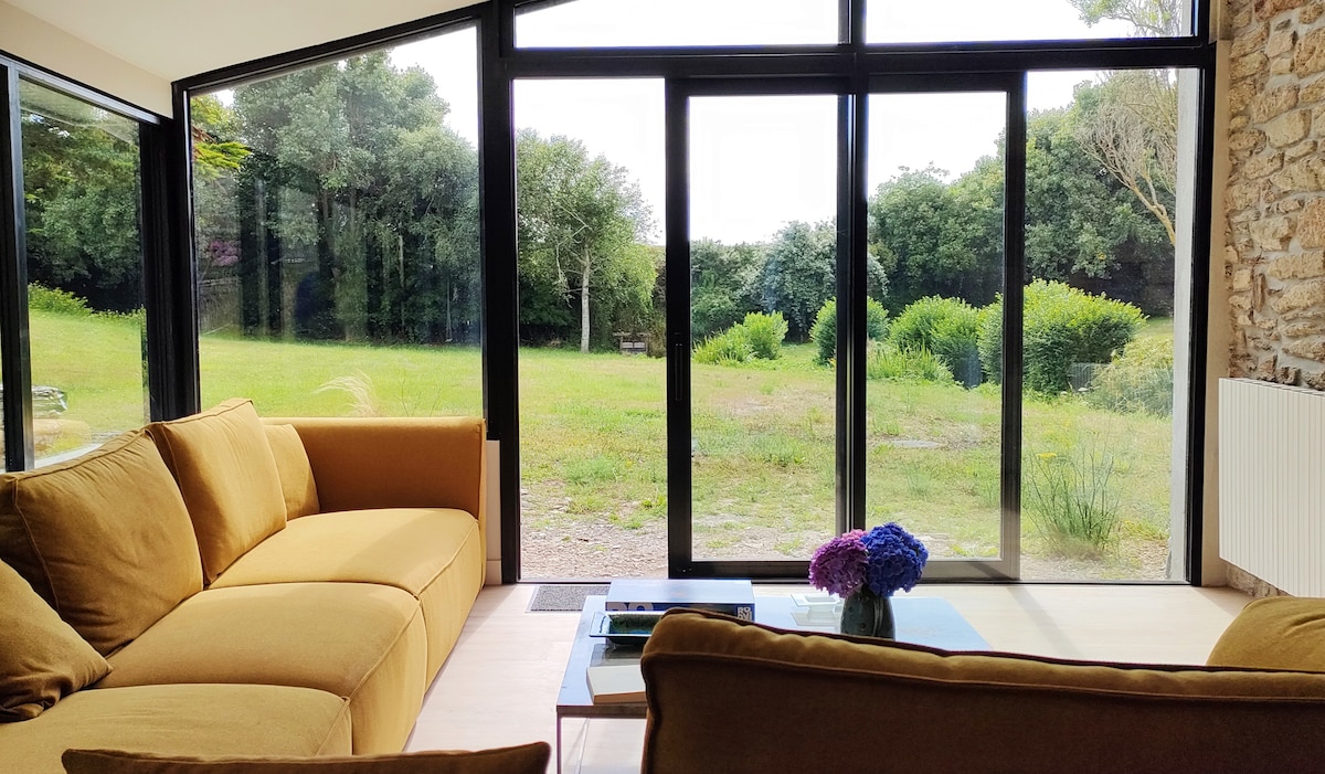 A bright and airy living space features a large yellow sofa facing expansive glass doors. Natural light floods the area, providing a view of the lush green garden outside, while a coffee table adorned with flowers adds a touch of color.