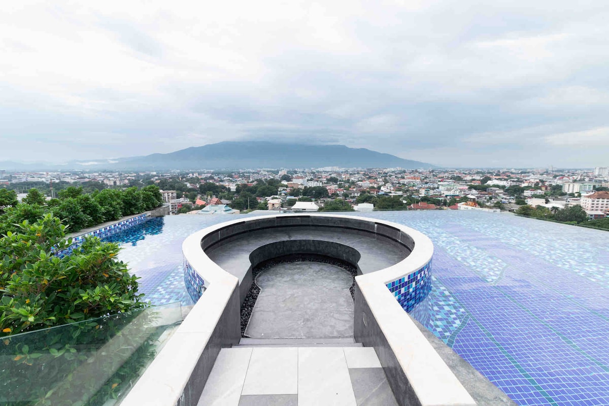 A panoramic view from the infinity pool on the 17th floor reveals the Chiang Mai cityscape. An expansive horizon is framed by distant mountains, while the water's edge creates a seamless transition into the skyline. Lush greenery borders the pool, enhancing the serene setting.