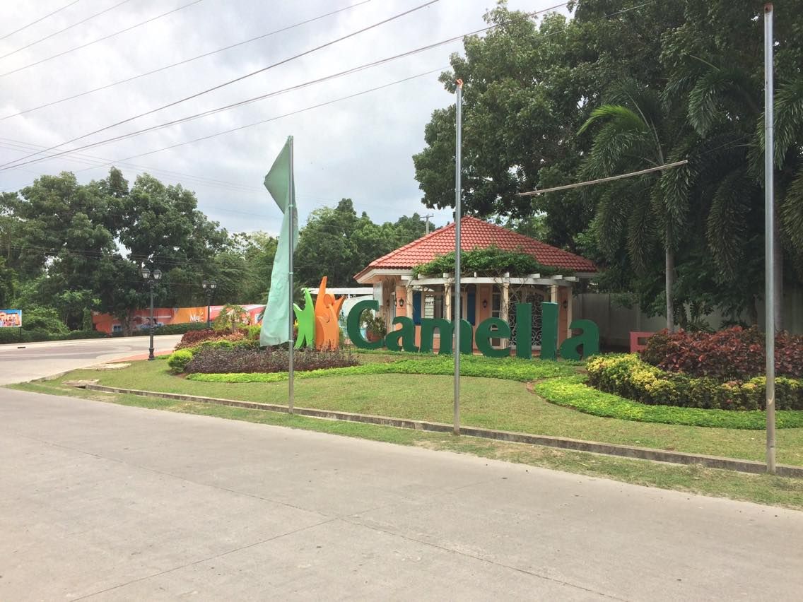The entrance of Camella Homes is marked by a green sign featuring the name 'Camella' prominently displayed. The landscaping includes well-maintained grass and trees, with a covered structure in the background topped with a red tile roof.