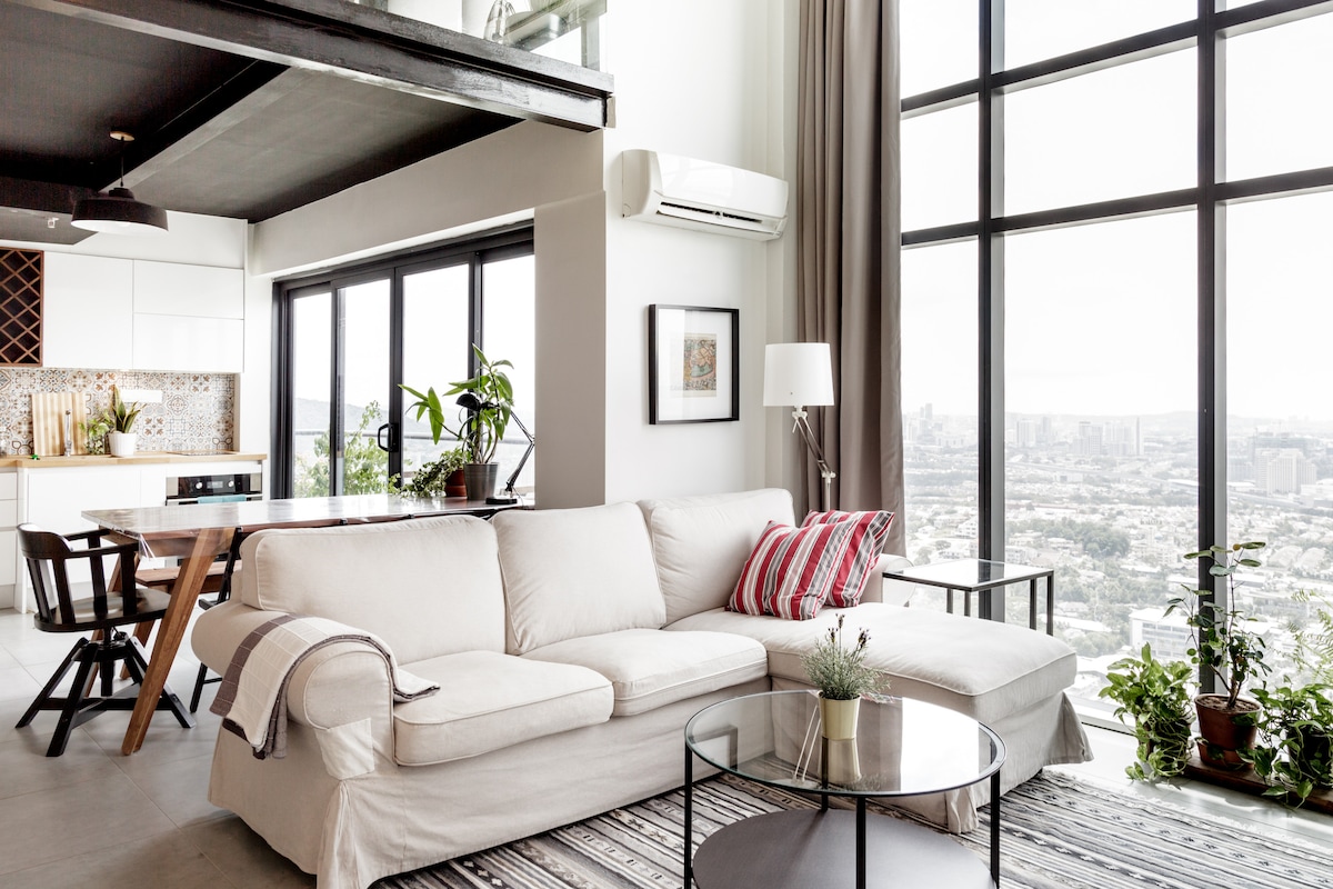 A bright and airy living space is highlighted by large windows offering expansive views of the city. A light-colored sectional sofa is placed in front of a glass coffee table, alongside potted plants. The open kitchen features modern finishes and pendant lighting above.