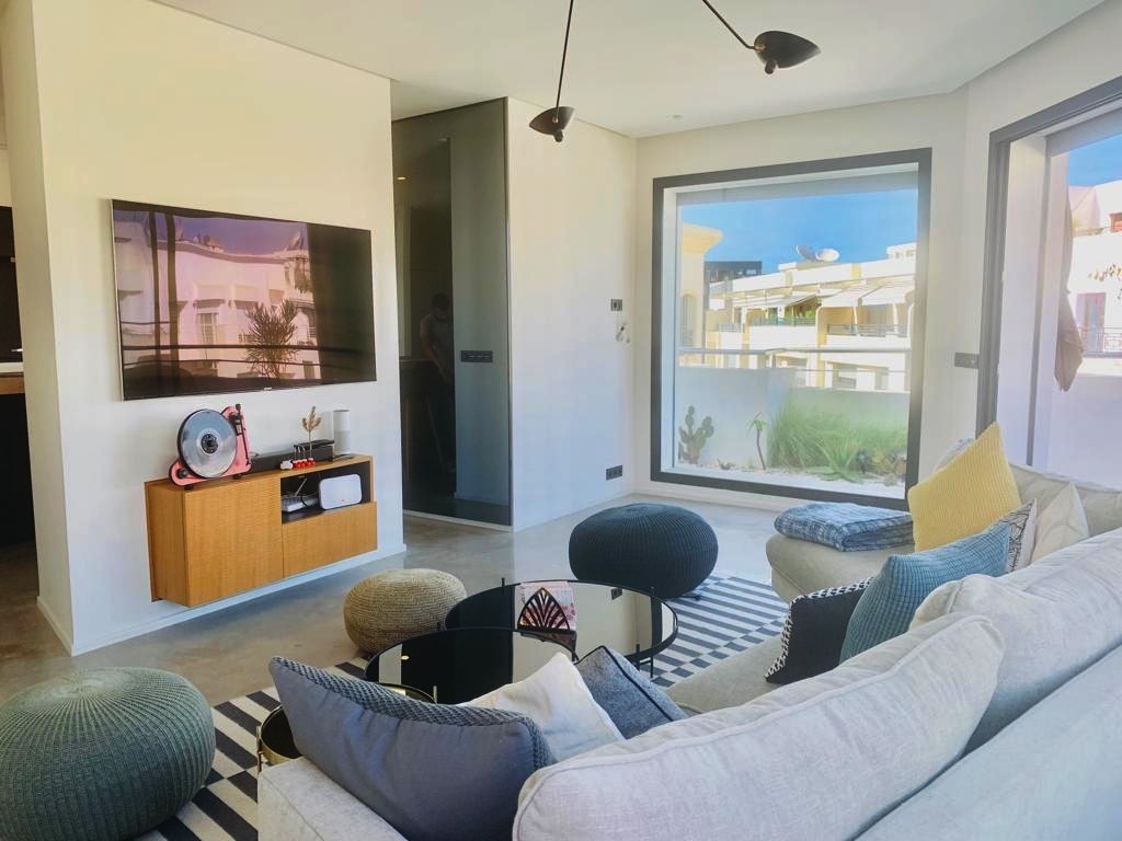 A modern living area is displayed, featuring a comfortable sofa adorned with various cushions. A coffee table is positioned in front of the seating, while a sleek TV is mounted on the wall. Large windows provide ample natural light and views of the outdoors.