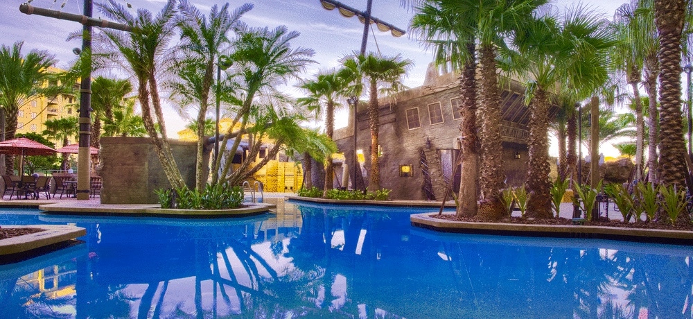 A vibrant pool area is framed by tropical palm trees, reflecting a playful pirate ship structure. The crystal-clear water invites relaxation, while lounge chairs with umbrellas are arranged around the pool, offering shade and seating for guests.