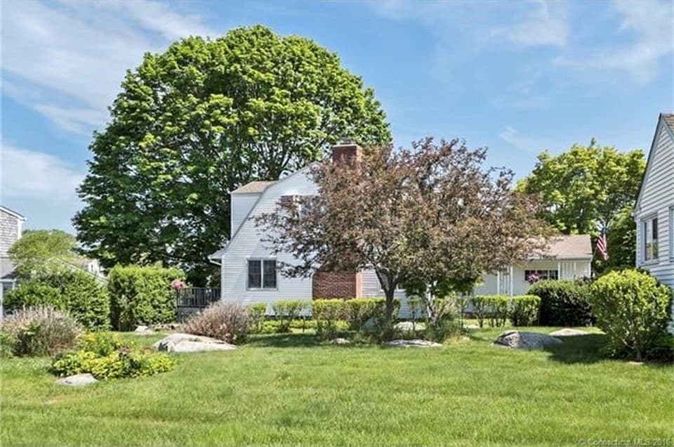 A well-maintained yard features a large green lawn surrounded by mature trees and shrubs. A prominent flowering tree adds a decorative touch, while smooth stones are scattered throughout the landscape, enhancing the natural beauty of the outdoor space.
