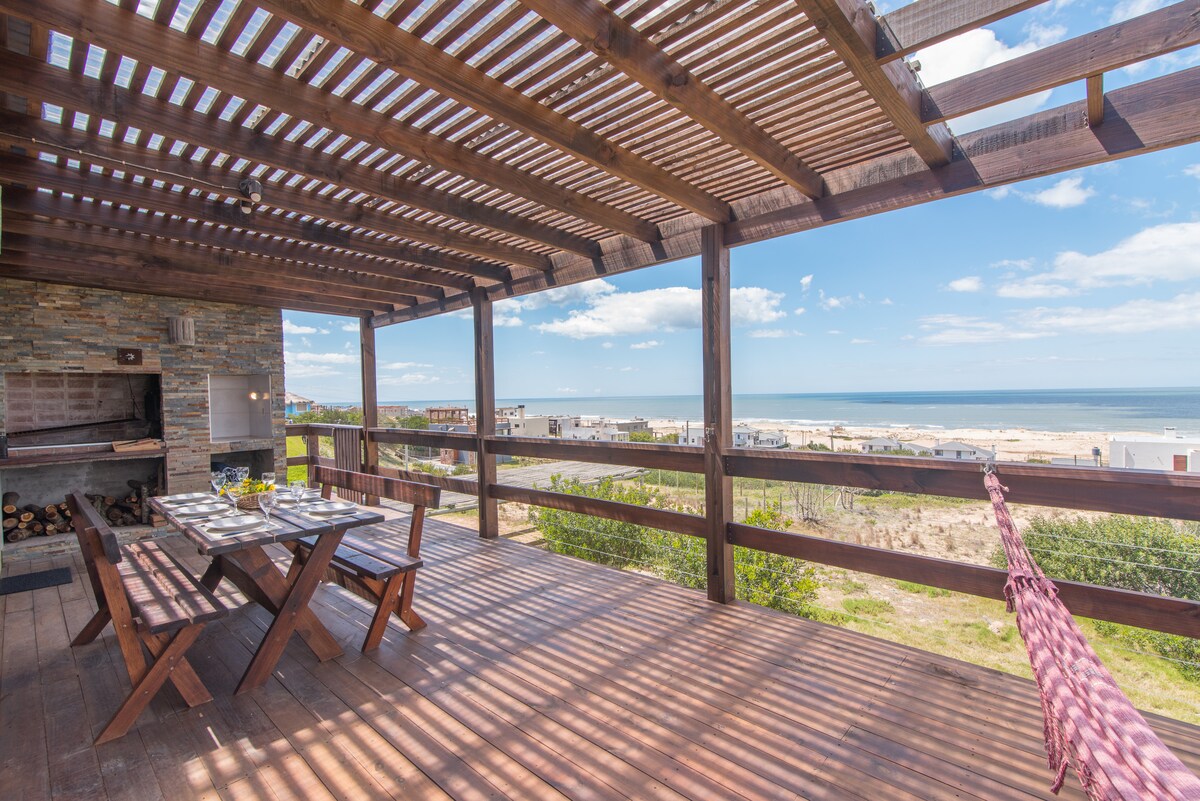 A spacious deck is depicted with a wooden table set for dining, complemented by a barbecue area. A hammock is visible, offering relaxation. The ocean view beyond is enhanced by natural light filtering through the slatted roofing, creating a soothing atmosphere.