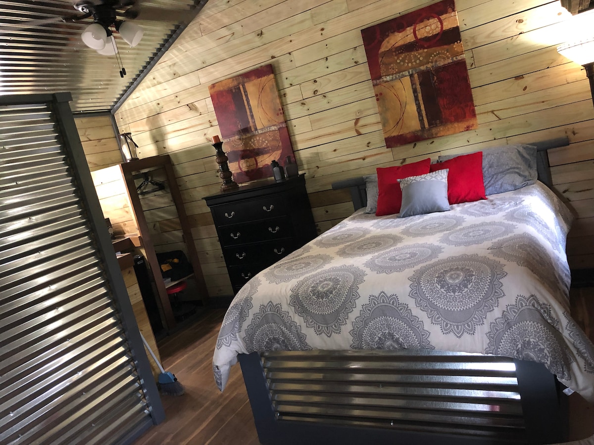 A rustic sleeping area features a spacious bed adorned with a patterned comforter, complemented by red decorative pillows. Simple artwork is hung above the bed, and a dark wooden dresser stands nearby, with wood-paneled walls and a polished floor enhancing the cabin's charm.