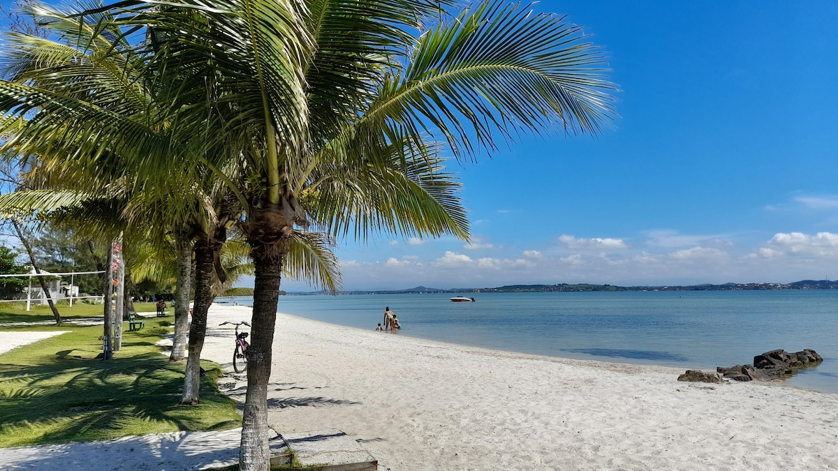 A sandy beach area is bordered by palm trees, providing shade and a tropical atmosphere. Calm waters extend to the horizon, with a few small boats visible in the distance. The scene is complemented by grassy areas and a playground nearby.