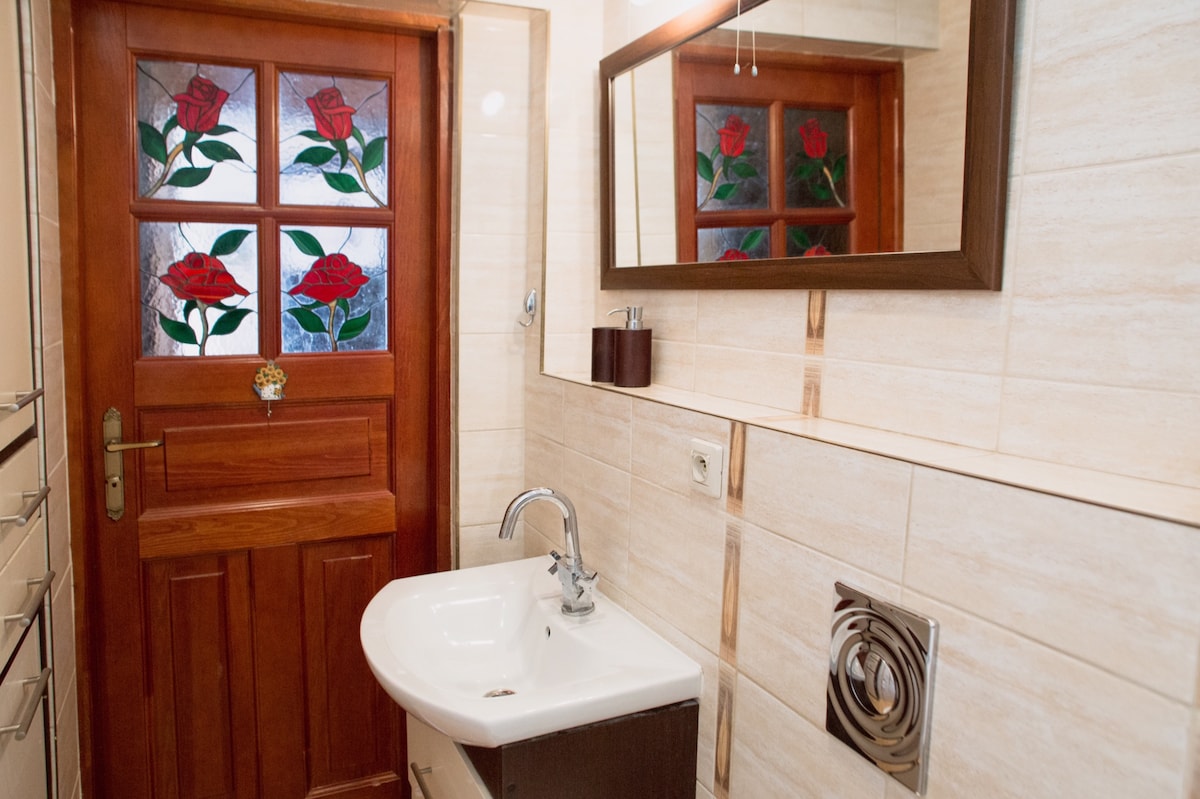 A well-appointed bathroom features a wooden door adorned with a stained glass panel depicting red roses. A white sink is mounted on a dark cabinet, and the walls are lined with neutral tiles. A wall mirror complements the overall design.