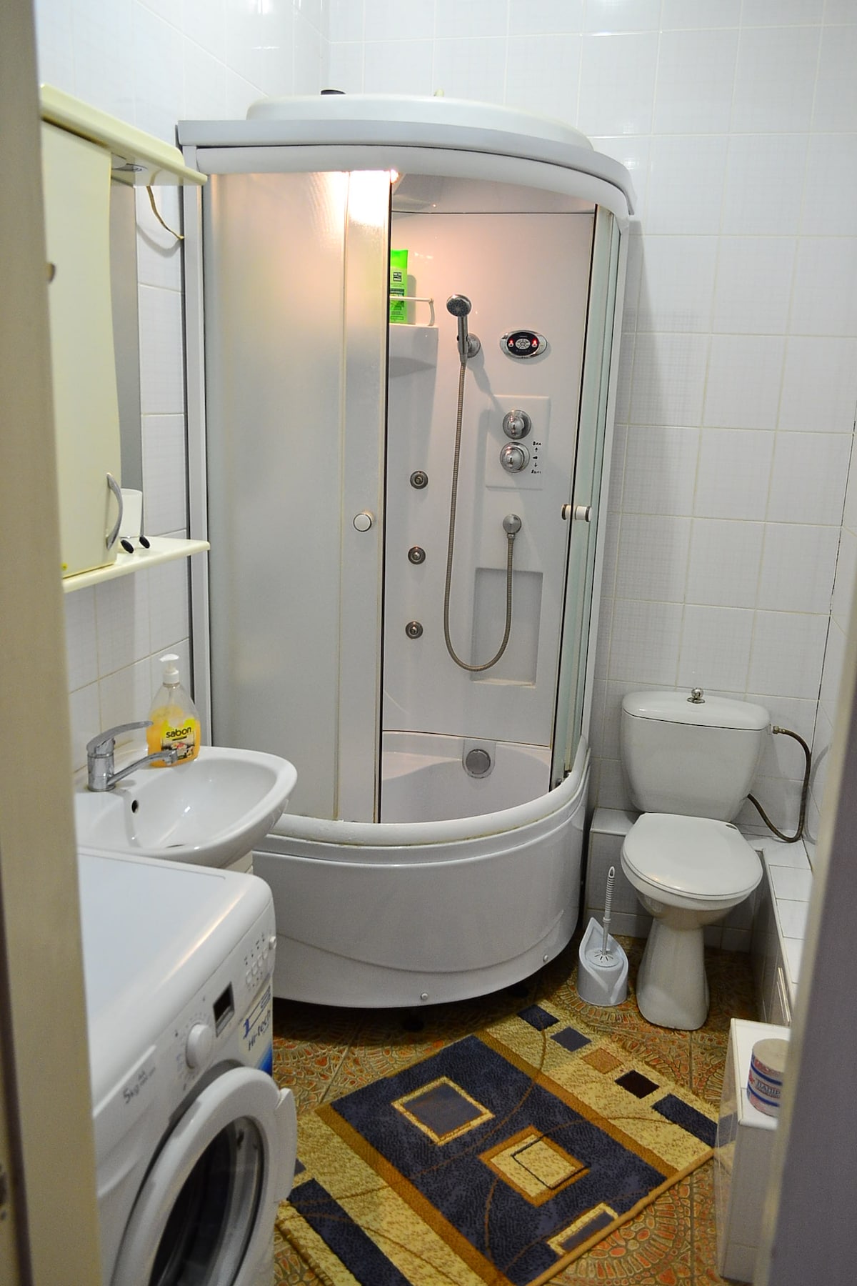 The bathroom features a transparent shower enclosure with an overhead and handheld shower system. A wash basin is positioned next to the shower, and a modern toilet is located beside it. The space is completed by a washing machine and tiled flooring.