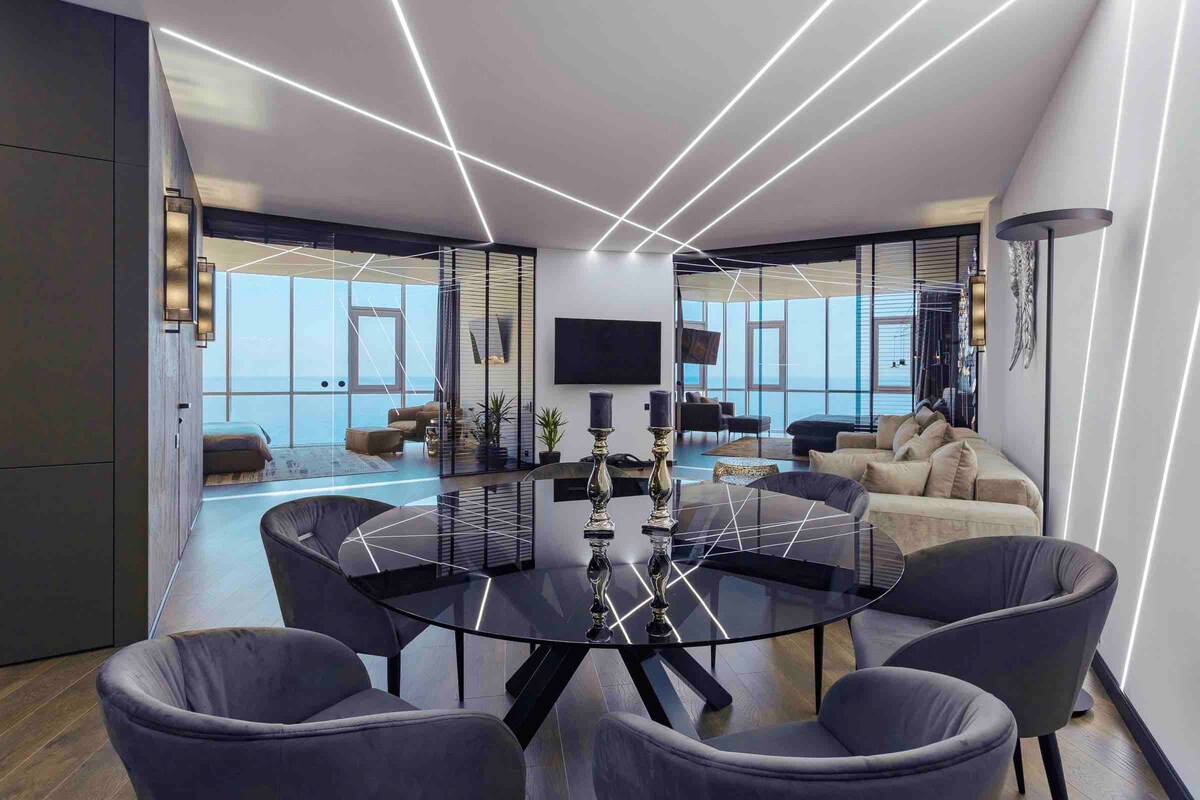The living area features a modern dining table surrounded by four plush chairs. A sleek entertainment center is positioned against the wall, with soft lighting accentuating the space. Floor-to-ceiling windows offer an expansive view of the sea, enhancing the bright and airy atmosphere.