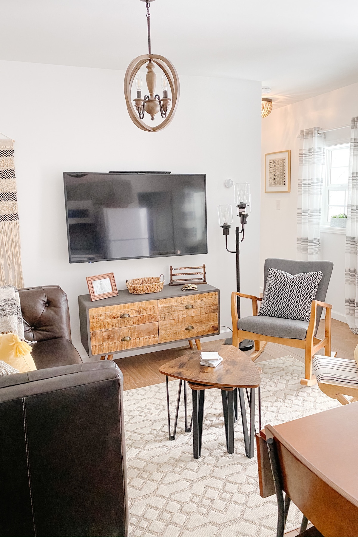 A cozy living area is presented with a television mounted on the wall. A black leather sofa faces a small wooden coffee table and two armchairs. Natural light filters through the windows, complemented by patterned curtains, creating a warm atmosphere.