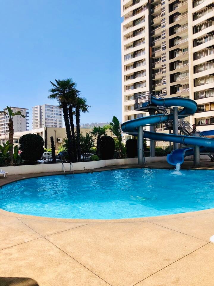 A refreshing pool is surrounded by lush greenery, featuring a fun water slide that cascades into the water. Tall buildings are visible in the background, under a clear blue sky, creating an inviting atmosphere for relaxation and enjoyment.