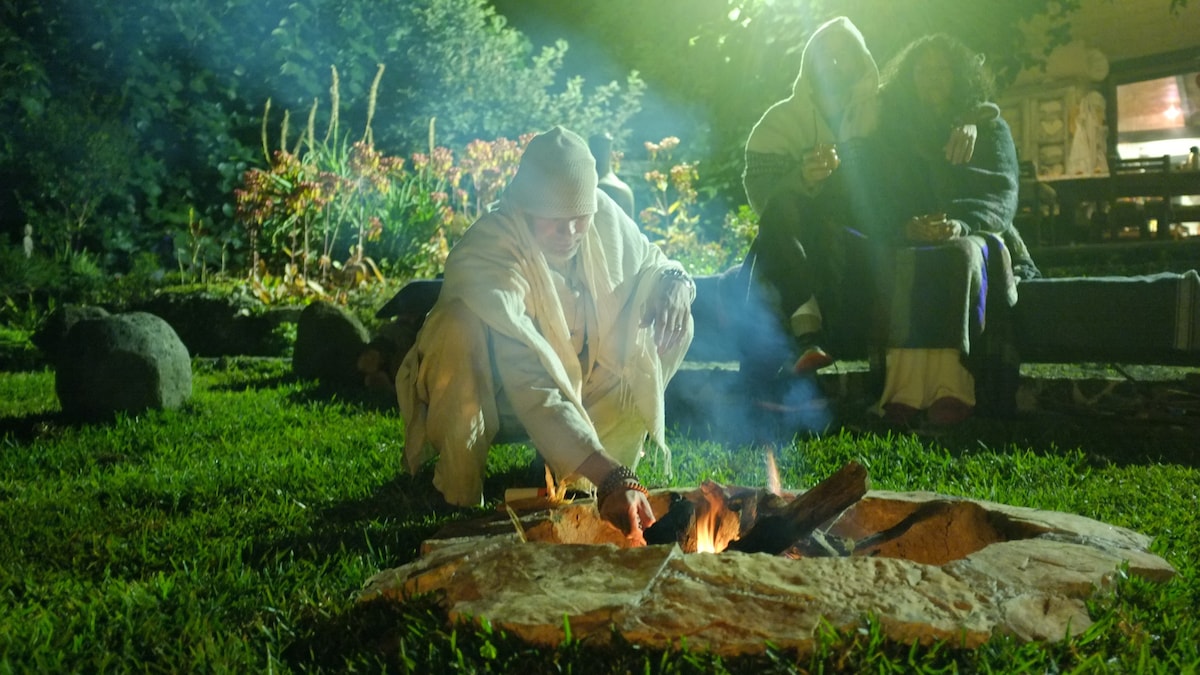 A cozy outdoor fire pit is surrounded by grassy terrain, where two individuals enjoy a relaxed conversation in the background. The fire flickers warmly, casting a gentle glow on nearby plants and creating a serene atmosphere under the night sky.