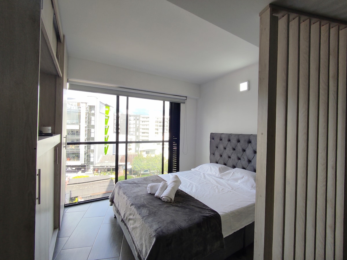 A modern bedroom features a spacious bed with neutral-toned bedding, framed by large floor-to-ceiling windows offering natural light and an urban view. Soft textures are observed with a gray throw and towels neatly placed at the foot of the bed. A minimalist wooden wardrobe is visible on one side.