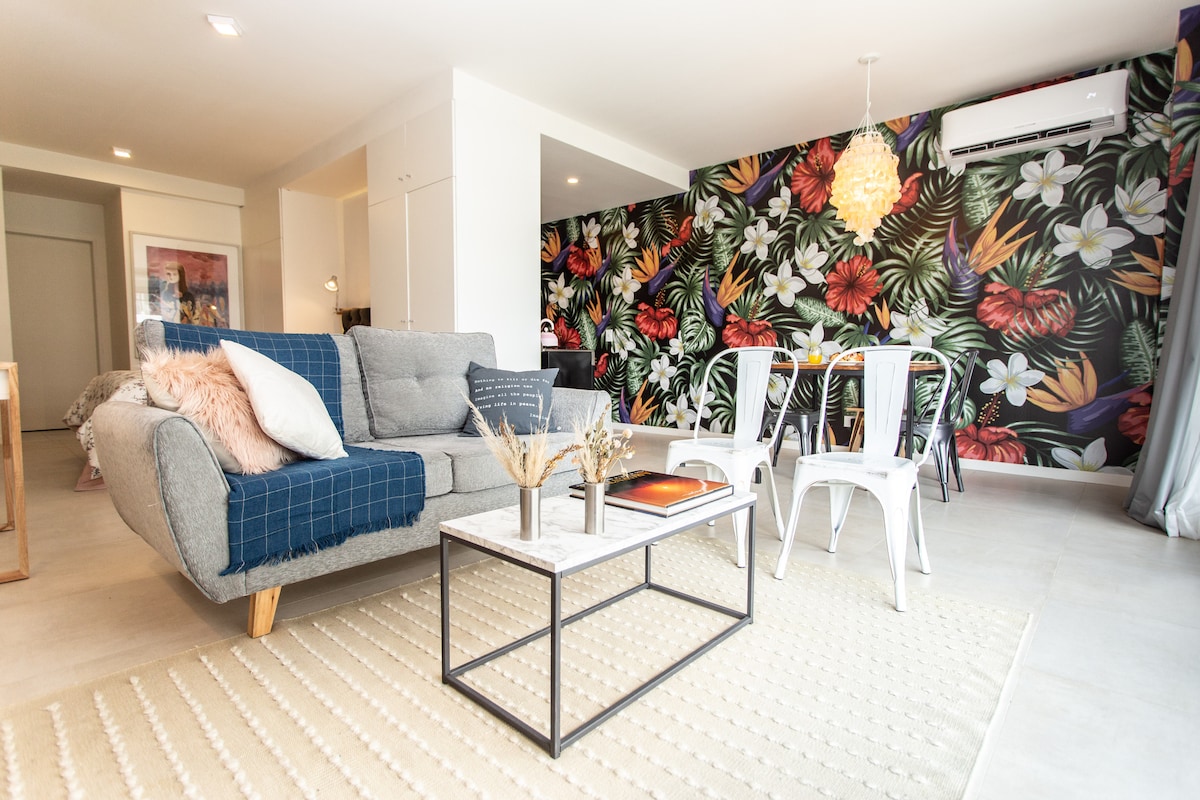 A cozy living area features a light-colored sofa adorned with soft cushions, complemented by a stylish coffee table with decorative elements. A vibrant floral wallpaper adds a lively touch to the space. A dining table with metal chairs is visible in the background.