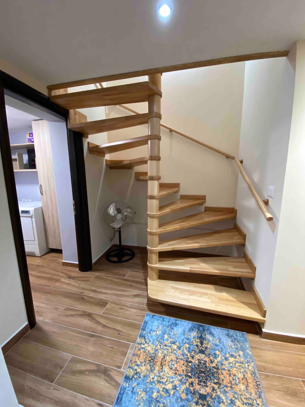 A spiral wooden staircase winds elegantly to the upper level, complemented by a small fan at its base. A textured blue rug adds a touch of color to the entryway, which is illuminated by a ceiling light. The surrounding walls are painted in soft neutral tones.