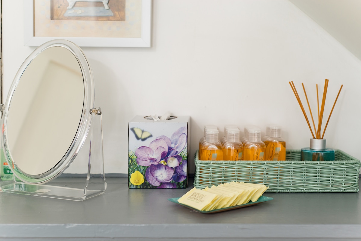 A well-organized surface displays a circular mirror and a decorative tissue box featuring purple flowers. Nearby, clear bottles are neatly arranged in a green wicker tray, accompanied by a small metallic container and a stack of paper coasters.