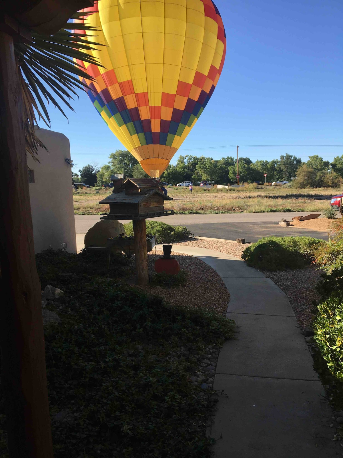 A colorful hot air balloon is seen hovering in the sky, featuring bright yellow and red patterns. A winding pathway is visible in the foreground, bordered by shrubs and boulders. The landscape includes open fields in the background, with a clear blue sky above.