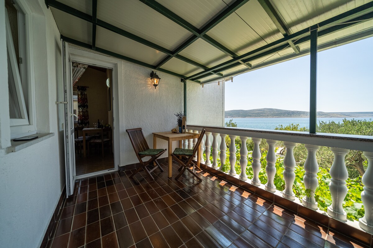 A covered terrace is presented with a wooden table and two chairs. The railing features classic white columns, providing a view of the glistening sea in the background. Sunlight filters gently through the open space, enhancing the serene environment.
