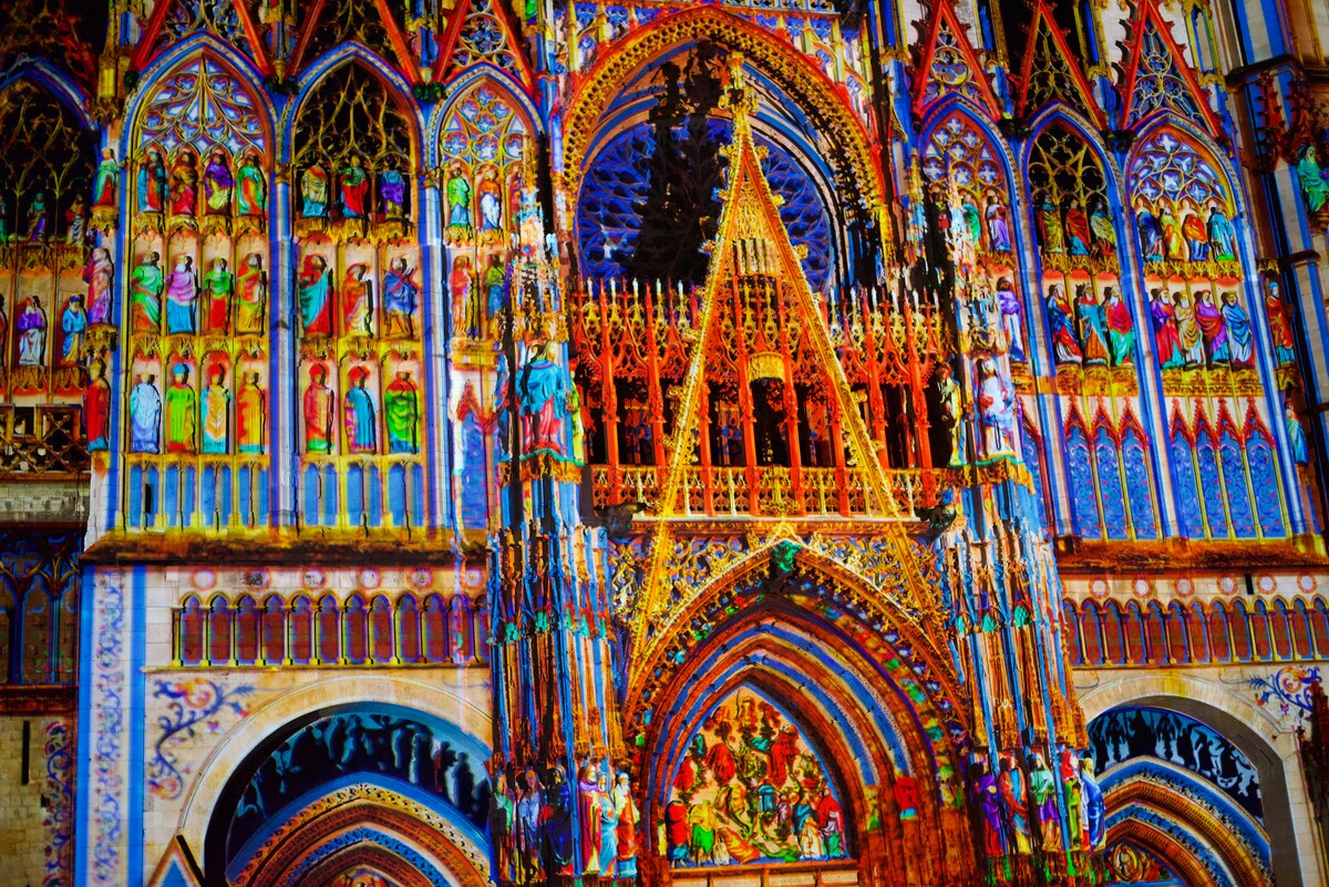A vibrant depiction of intricate architectural details is displayed on a cathedral facade. Colorful projections highlight numerous figures and ornate decorations, showcasing the artistry of the structure. Elements such as arches, statues, and elaborate wooden carvings are prominently featured.