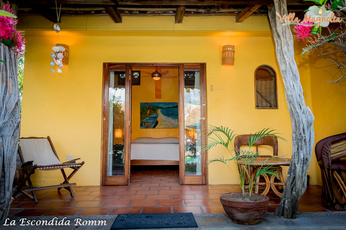 The entrance to La Escondida room features rustic wooden doors, framed by vibrant yellow walls. A comfortable seating area with decorative plants is visible, alongside two patio chairs. Inside, a spacious bed is highlighted by colorful wall art, welcoming natural light through the open doorway.