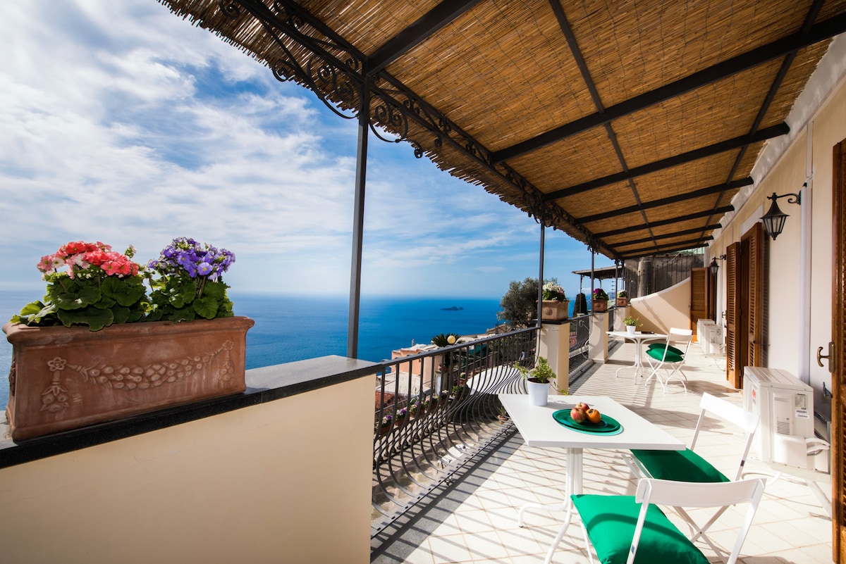 The spacious terrace features a thatched overhang providing shade, enhanced by colorful planters with blooming flowers. Outdoor furniture, including tables and chairs, faces unobstructed views of the tranquil blue sea and sky, creating an inviting space for relaxation and dining.