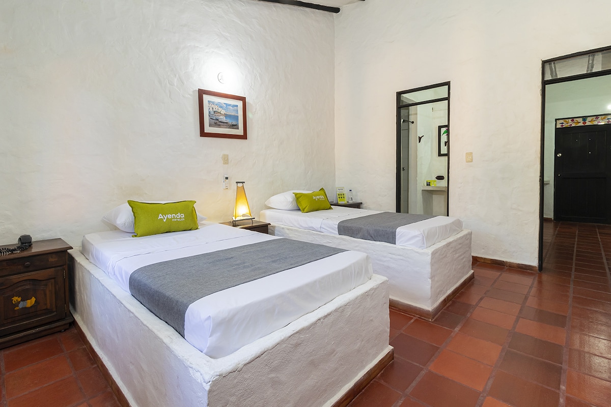 The room features two spacious beds with gray linens, positioned against textured white walls. A bedside table holds a lamp, while a decorative painting complements the space. An en-suite bathroom is visible through an open door, enhancing the room's functionality.