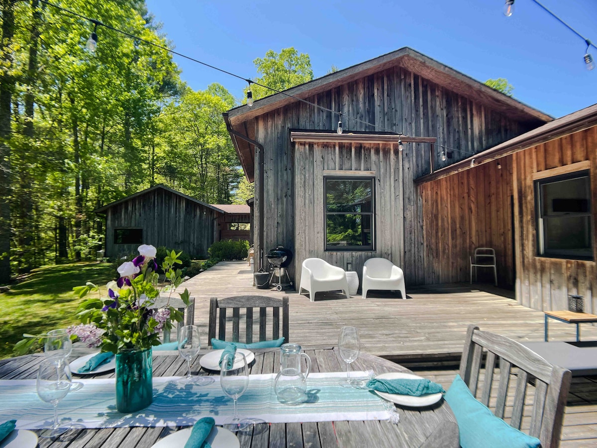 A charming outdoor dining area is set with a wooden table and glassware, centered in front of a rustic cabin. The background features a wooden structure with large windows, surrounded by lush greenery and soft sunlight filtering through the trees.