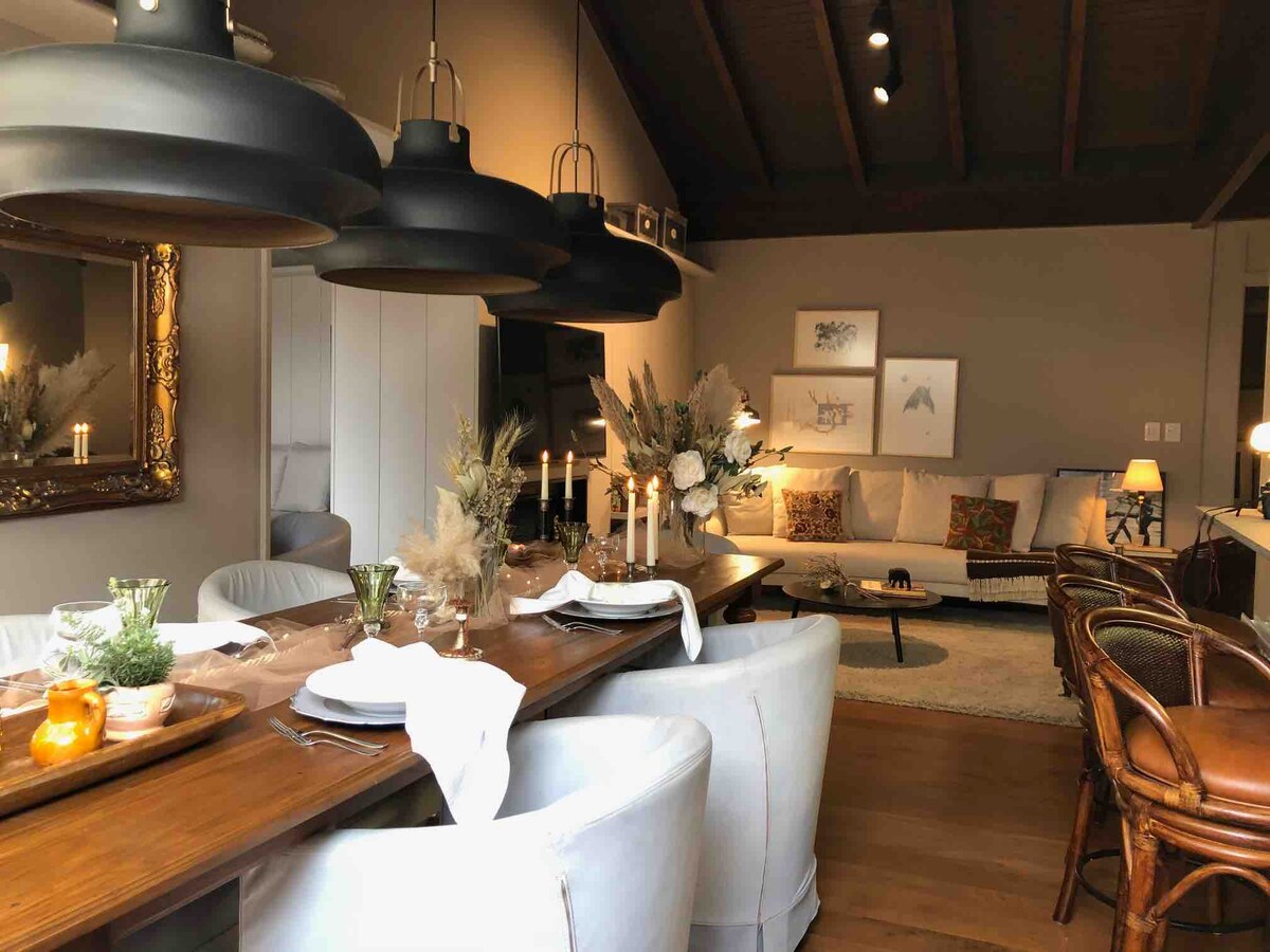 An elegant dining area is showcased, featuring a long wooden table set with plates and glassware. Soft, ambient lighting is provided by large pendant lamps. A cozy living space is seen in the background, accented by comfortable seating and decorative artwork.