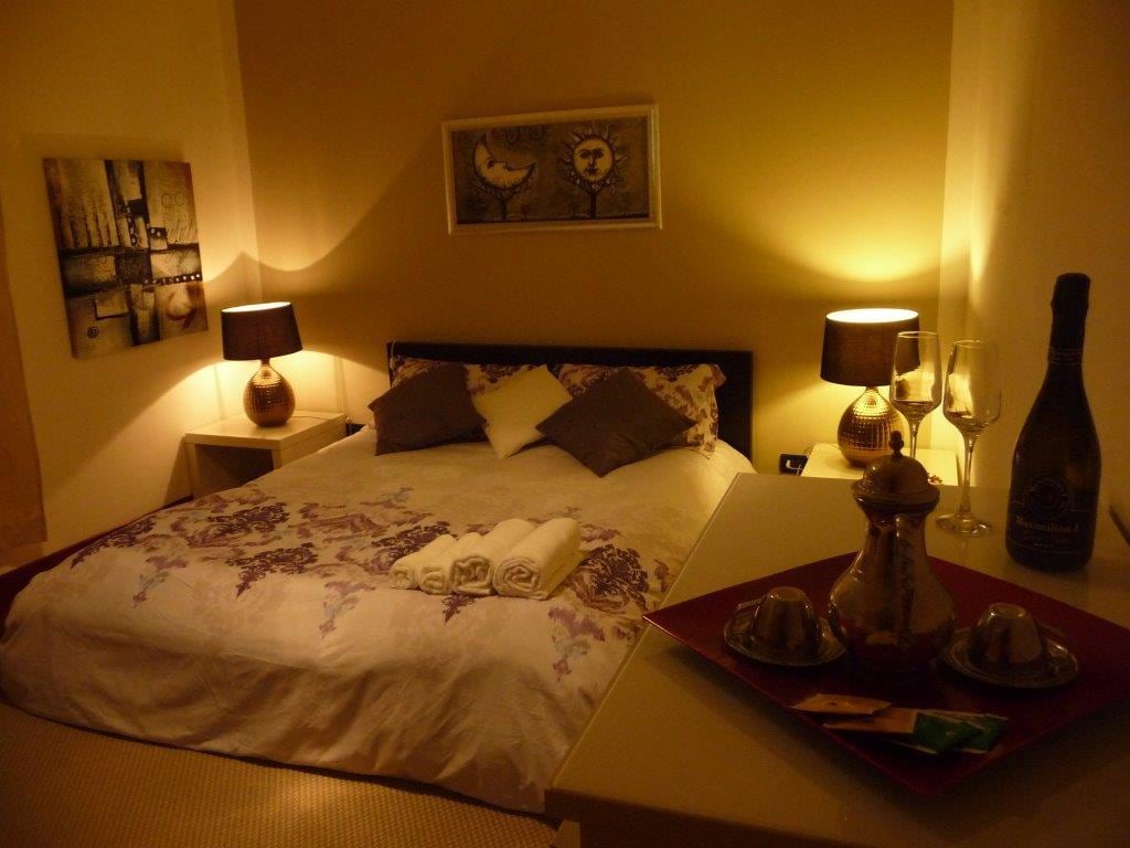 A well-lit bedroom features a spacious bed adorned with decorative pillows and a soft blanket. Two bedside lamps provide a warm glow, complementing a wall art piece above the bed. A small table displays elegant glassware and a bottle, setting a cozy ambiance.